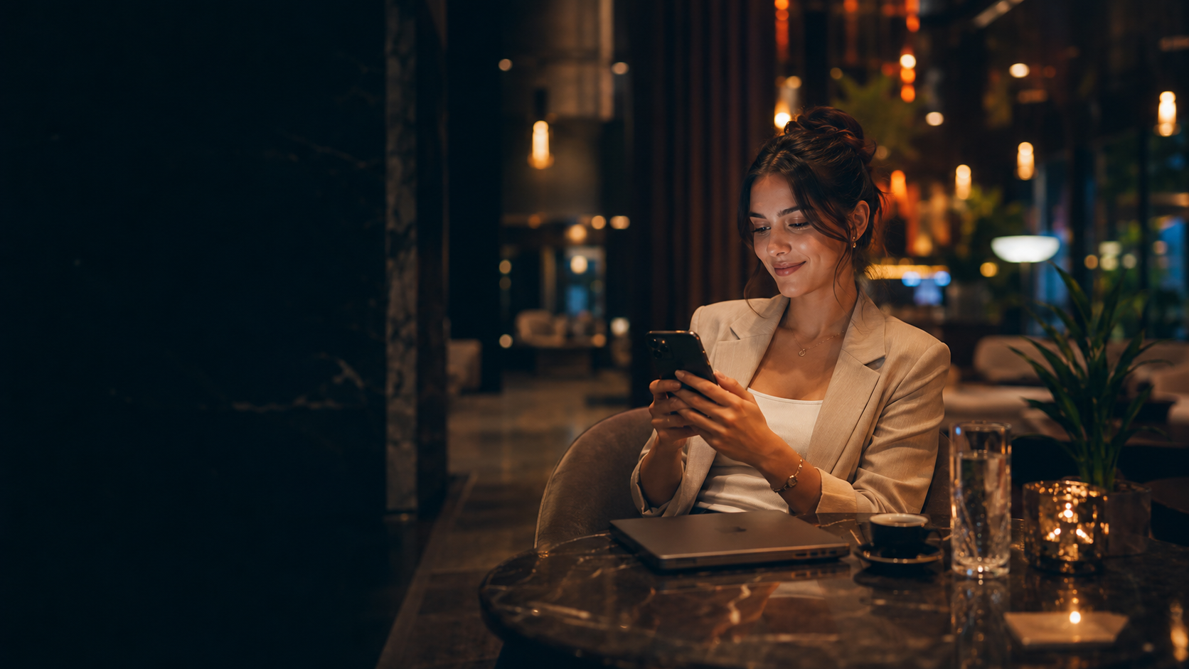 Woman smiling at her phone in a warm candle-lit café, city lights glowing behind her